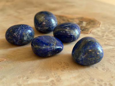 Pierres roulées en lapis-lazuli naturel de qualité A, aux nuances de bleu profond parsemées de reflets dorés, disposées sur un fond en bois clair.